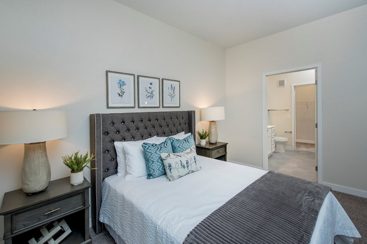 A modern bedroom with stylish decor, featuring a bed with patterned pillows, framed artwork above the headboard, nightstands with lamps, and potted plants.
