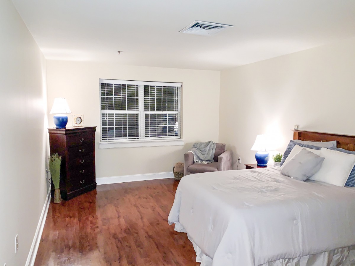 A cozy bedroom with a bed, armchair, and wooden dresser, featuring a warm hardwood floor and soft lighting.