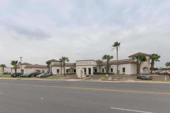 A white building with palm trees lining the entrance, situated along a road under a cloudy sky.