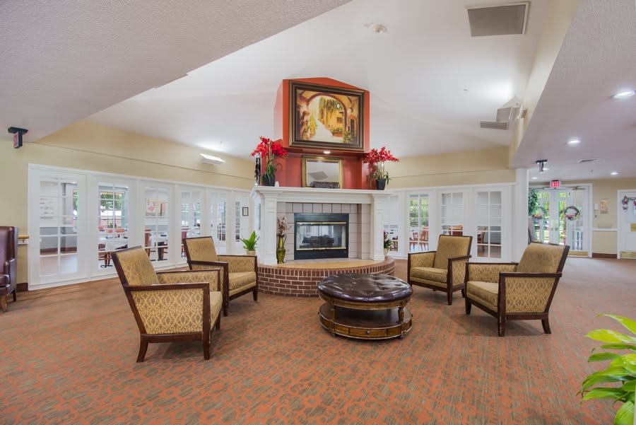 A cozy living room with a central fireplace, surrounded by comfortable chairs and a coffee table, featuring artwork and plants for decoration.