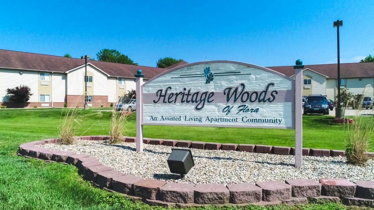 Sign for Heritage Woods, an assisted living apartment community, with buildings and a clear blue sky in the background.