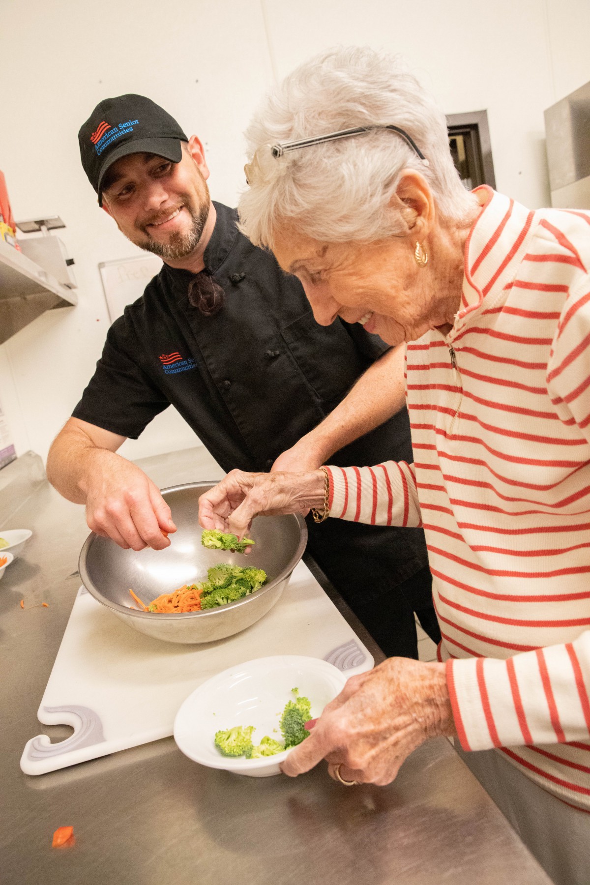 A chef assists an elderly person in preparing a salad in the kitchen.