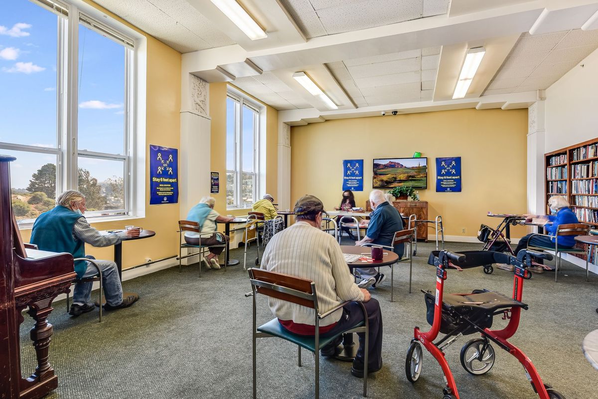 A community center room where several senior individuals are gathered, sitting at tables and socializing.