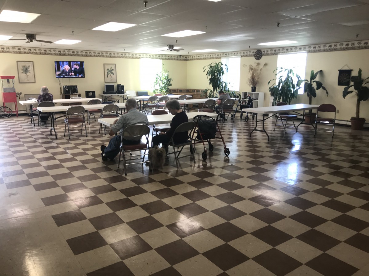A community center meeting room with tables and chairs arranged sparsely around the checkered floor, a few people seated.