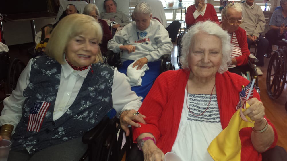 Two elderly women smiling and sitting in wheelchairs indoors.