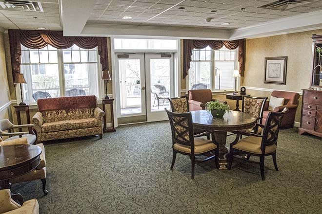 A cozy living room with a large table, chairs, and a sofa, featuring carpeted flooring and large windows with drapes.
