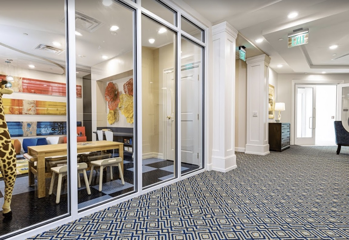 A modern office interior featuring a glass-walled conference room with artwork on the wall and patterned carpeting.