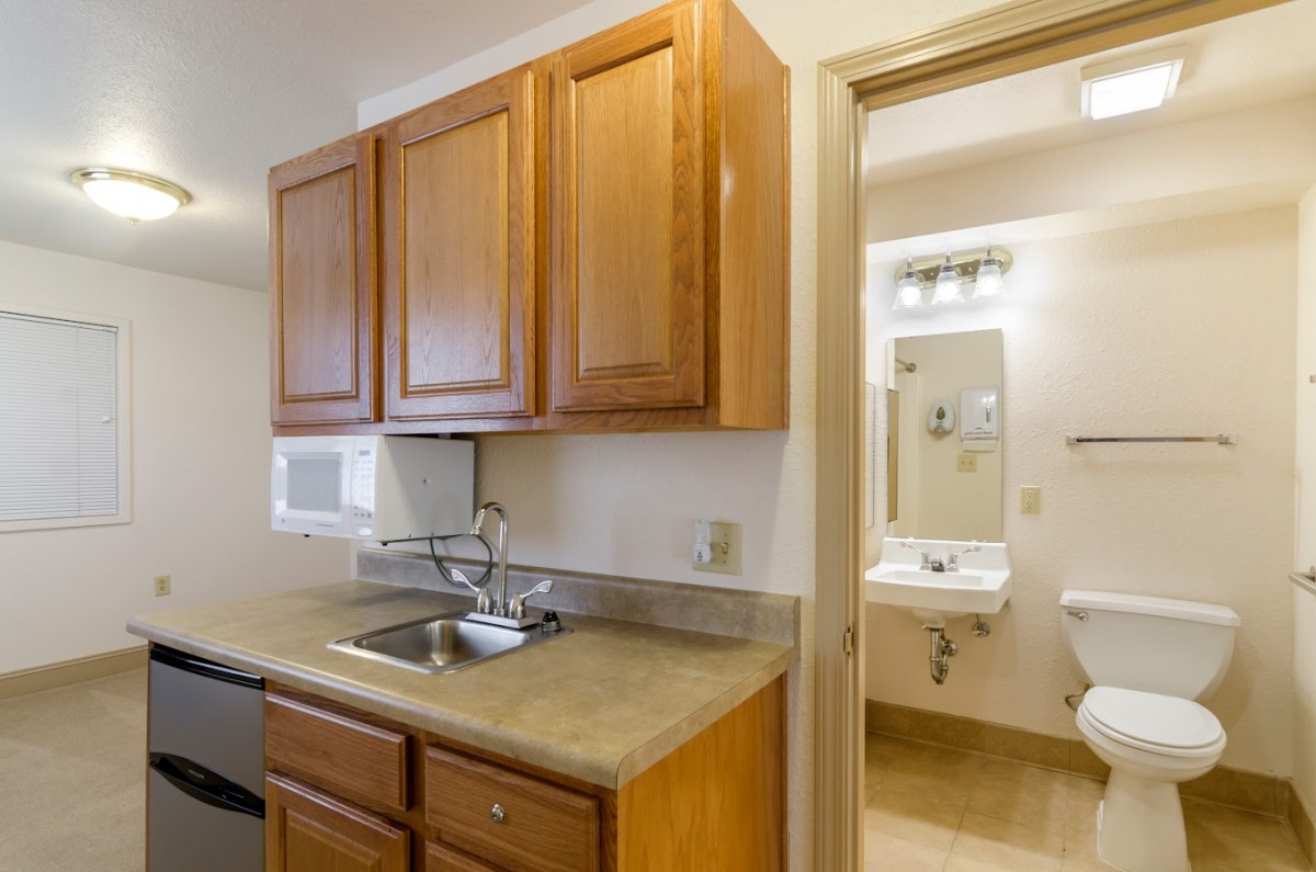 A small kitchen area with wooden cabinets, a sink, and a view into an adjacent bathroom.