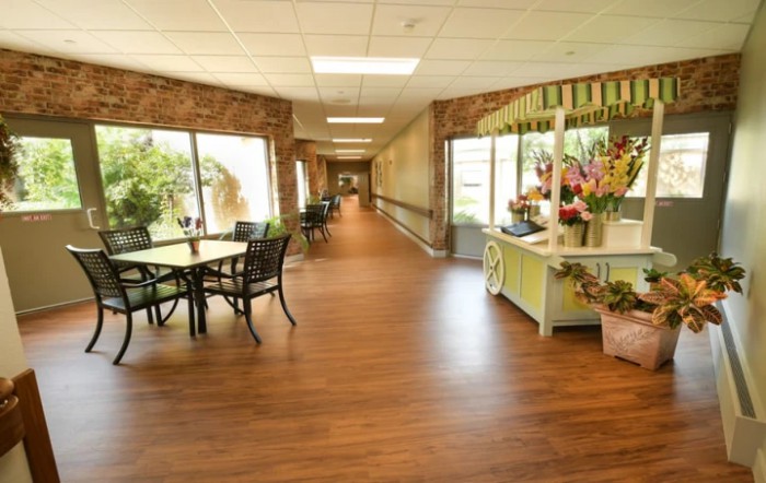 A bright hallway with wooden floors, tables, chairs, and decorative flowers along the side.