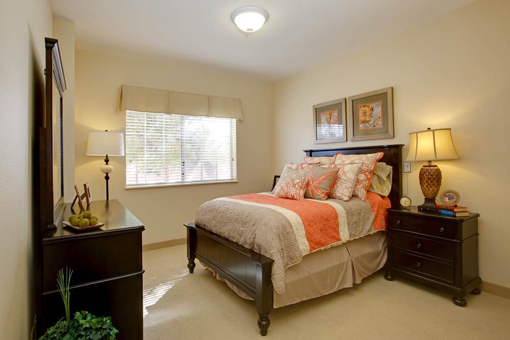 A cozy bedroom with a neatly made bed featuring orange and beige bedding, flanked by bedside tables with lamps, and artwork hanging above the bed.