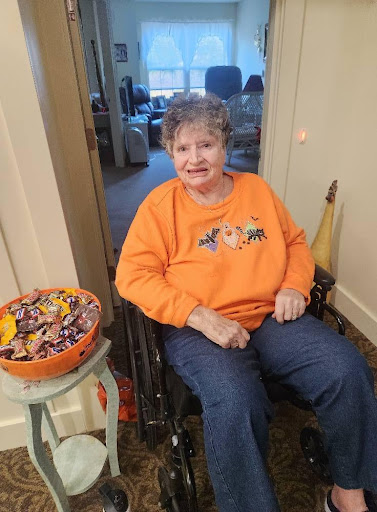 A smiling woman in a wheelchair, dressed in an orange sweater for Halloween, sitting beside a bowl of candy.