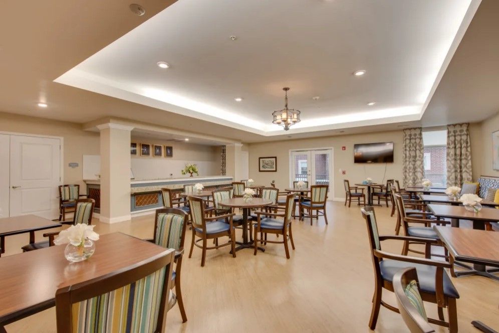 A well-lit dining area with neatly arranged tables and chairs, featuring a central chandelier and modern decor.