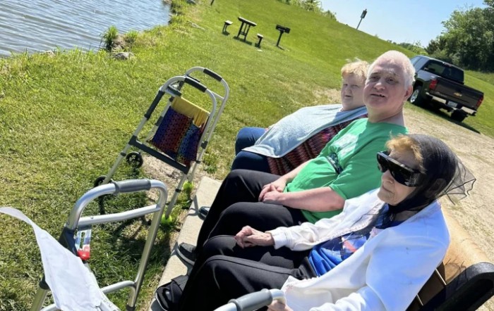 A group of people enjoying a sunny day at the park, seated near the water with a walker nearby.