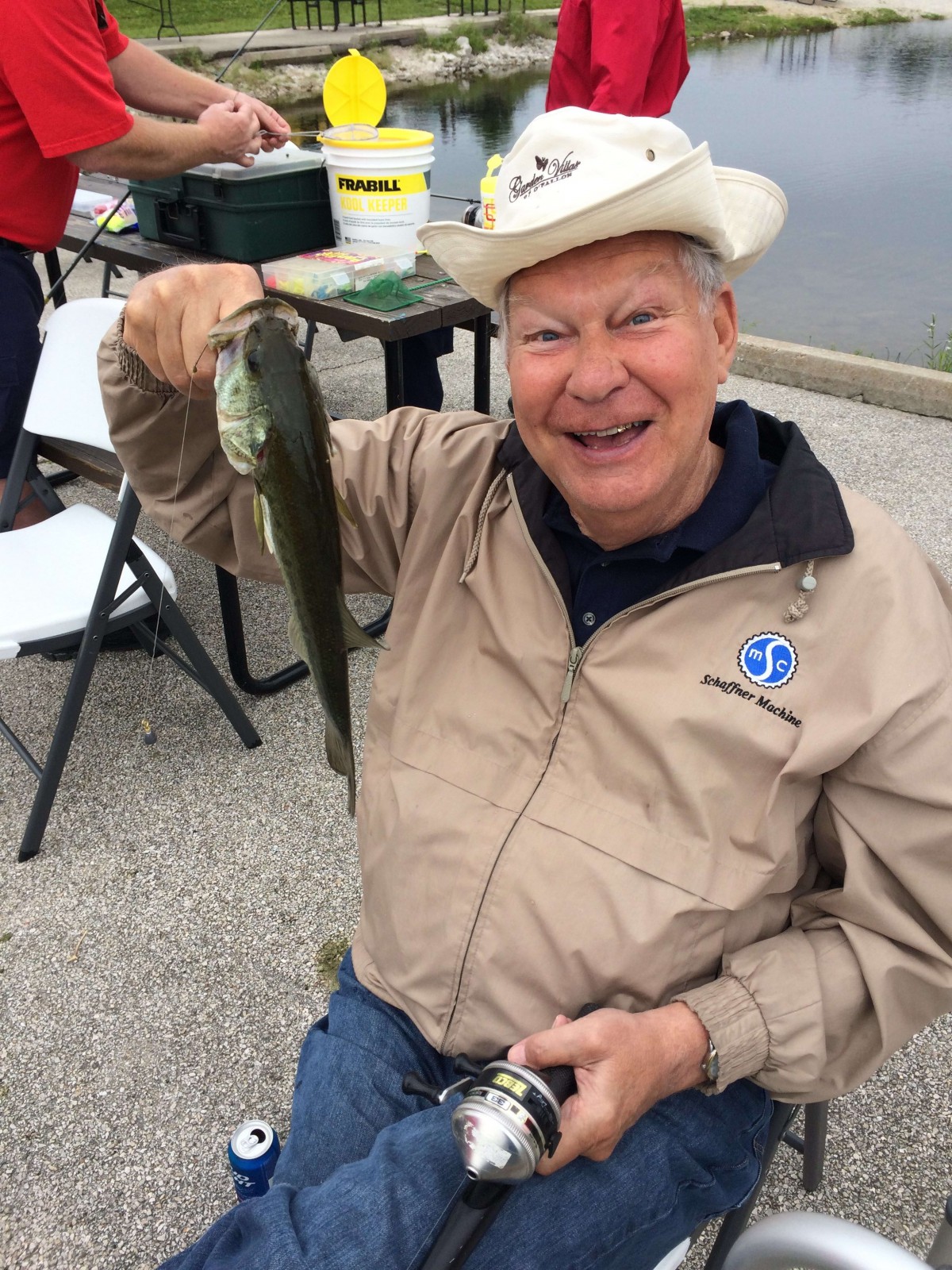 A smiling man holding a fish while sitting outdoors, wearing a hat and casual jacket.