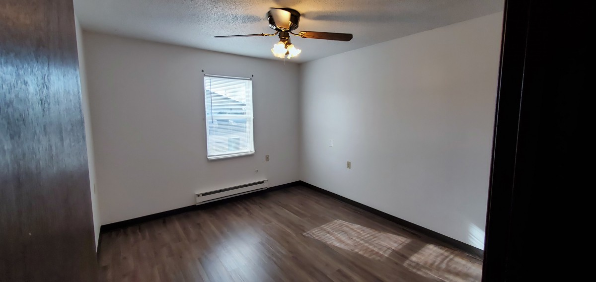 An empty room with hardwood flooring, a ceiling fan, and a window allowing natural light.