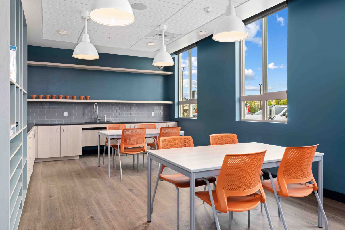 A modern office breakroom with tables and orange chairs under pendant lighting.