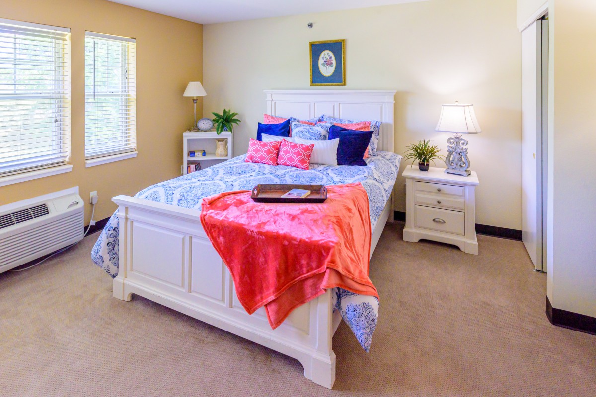 A cozy bedroom with a neatly made bed, colorful pillows, and a bright coral blanket. The room features a nightstand with a lamp, plants for decoration, and natural light coming through the window.