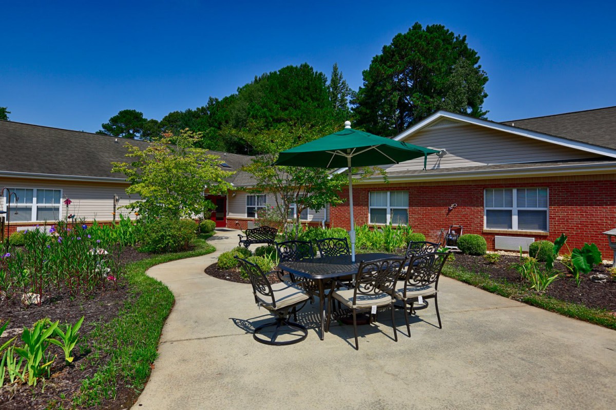 A sunny outdoor patio with a table, chairs, and an umbrella surrounded by a landscaped garden.