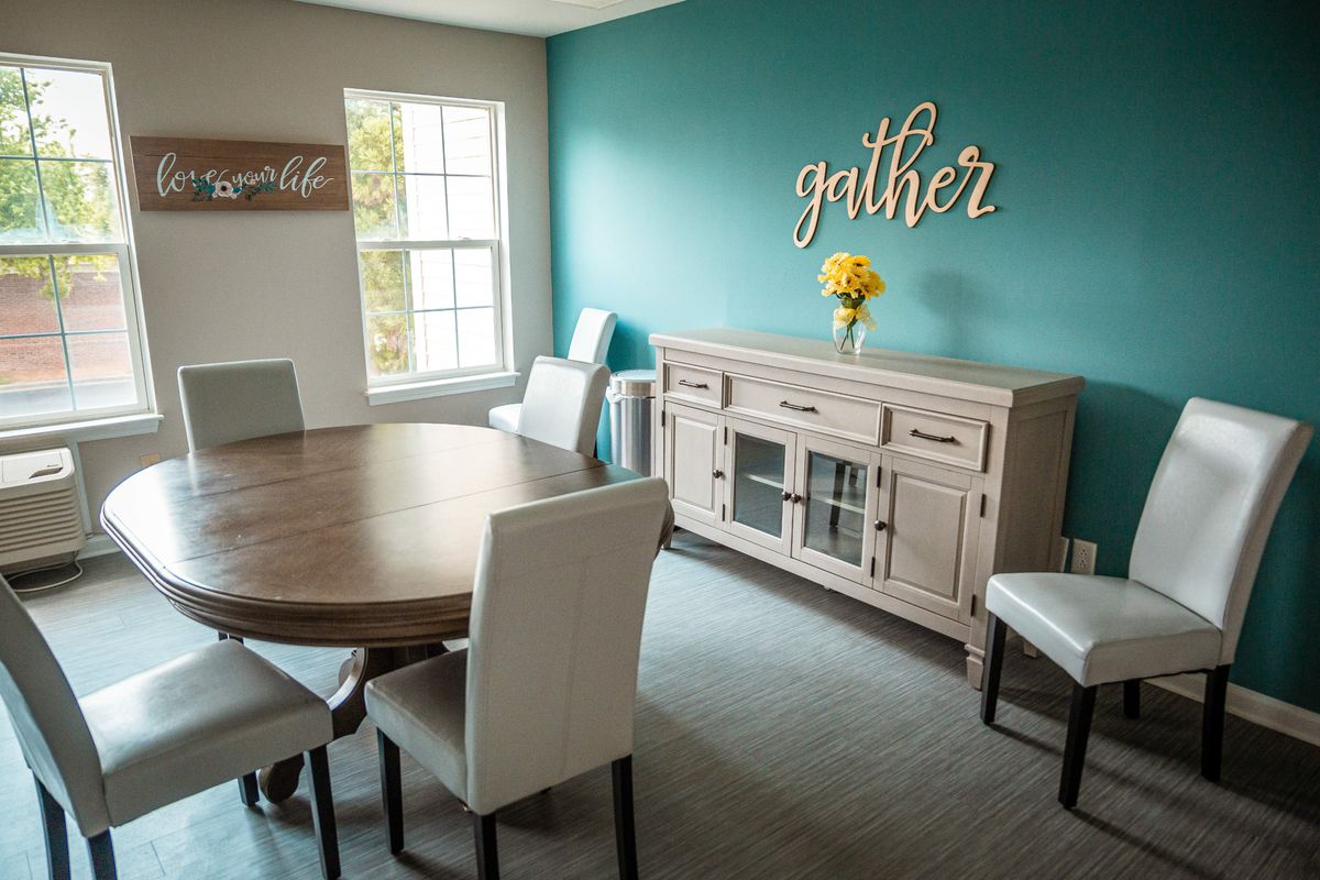 A modern dining room with a wooden table, upholstered chairs, and a sideboard, accented by wall art and a vase of yellow flowers.