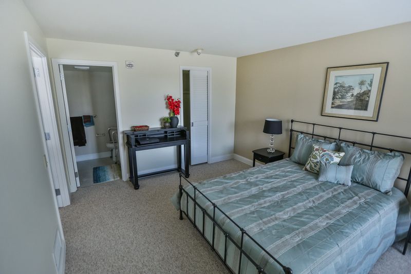 A modern bedroom featuring a bed with a striped comforter, adjacent nightstand with a lamp, and a desk with floral decorations near the bathroom entrance.