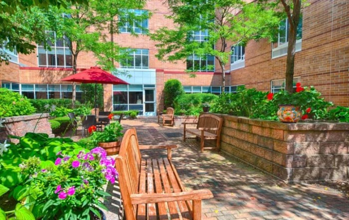 A tranquil garden patio area with wooden benches, colorful flowers, and lush greenery in front of a brick building.
