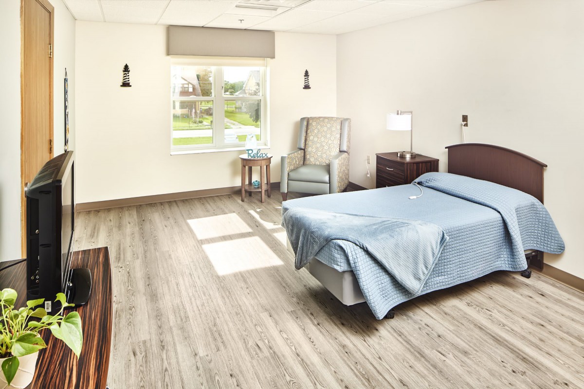 A clean and minimalist hospital room with a single bed, a chair, and a window letting in natural light.