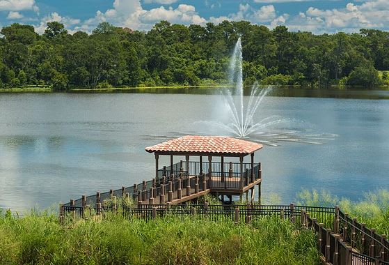 A scenic view of a gazebo by a lake with a fountain and lush greenery.