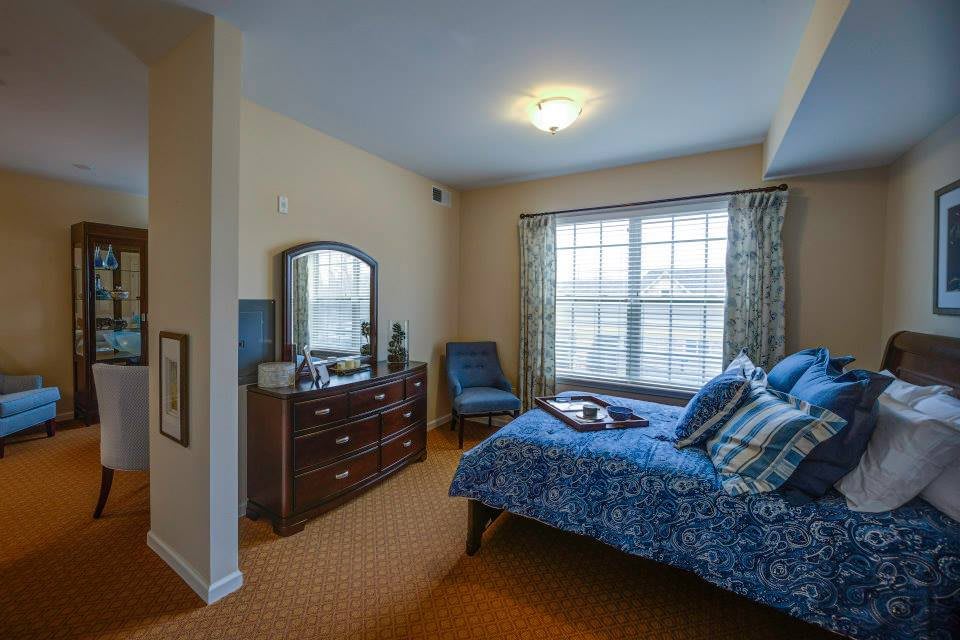 A cozy bedroom featuring a bed with blue bedding, a wooden dresser with a mirror, and a window with patterned curtains.