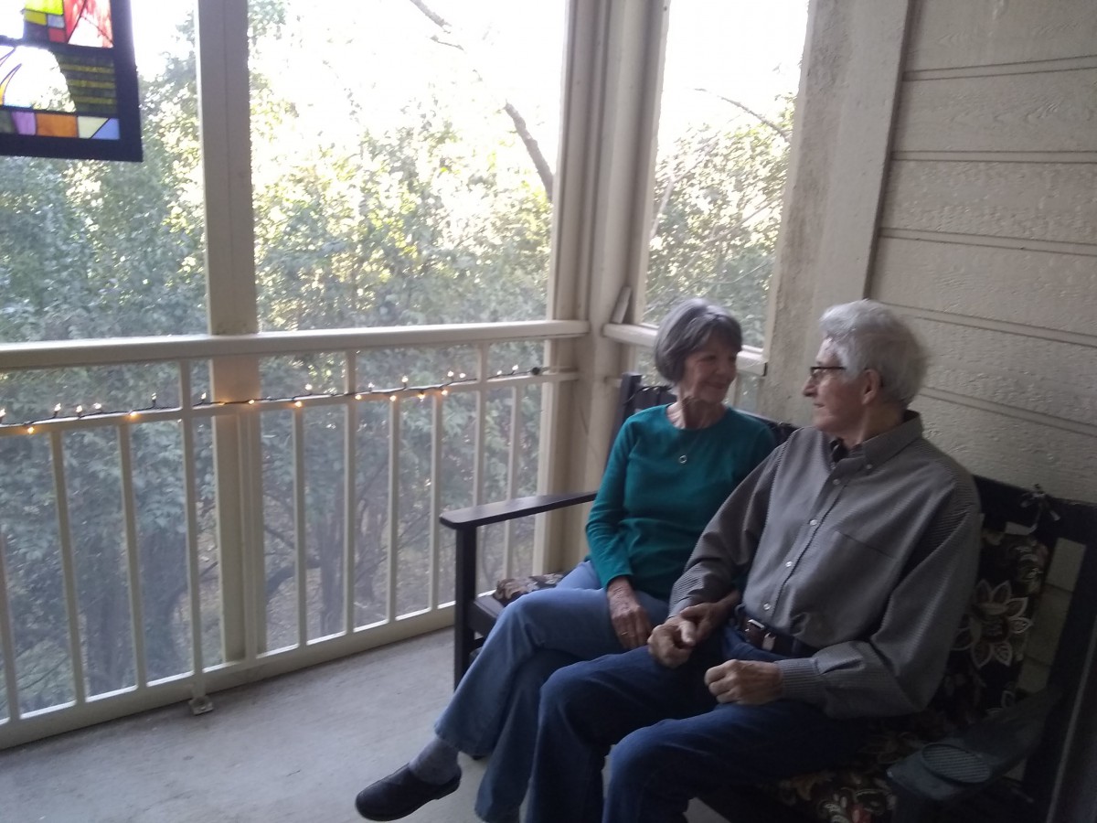 An elderly couple sits on a porch, enjoying a conversation in the daylight.