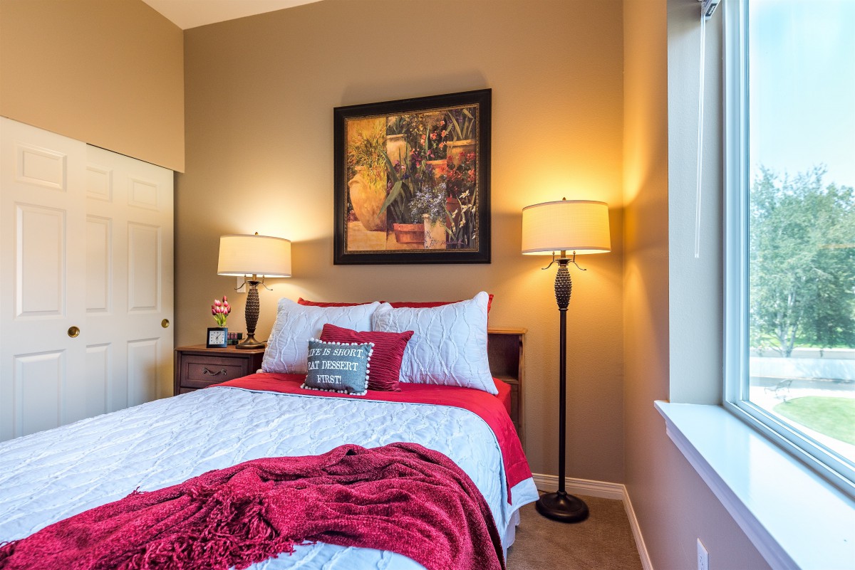 A cozy bedroom with a neatly made bed, featuring red and white bedding, flanked by two floor lamps and adorned with wall art.