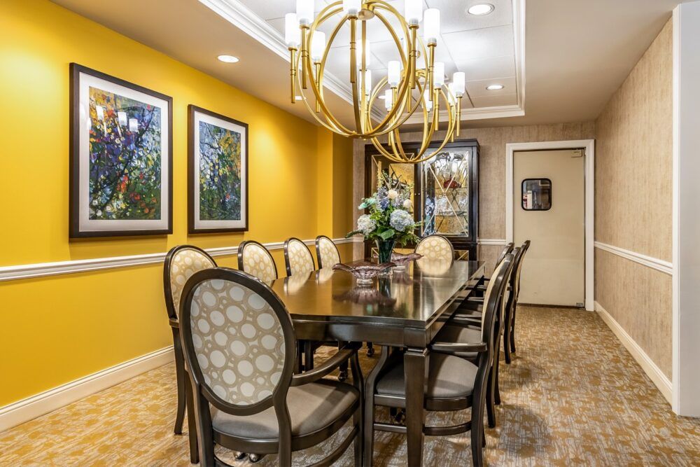 A stylish dining room featuring a large table with upholstered chairs, an elegant chandelier, and vibrant yellow walls adorned with artwork.