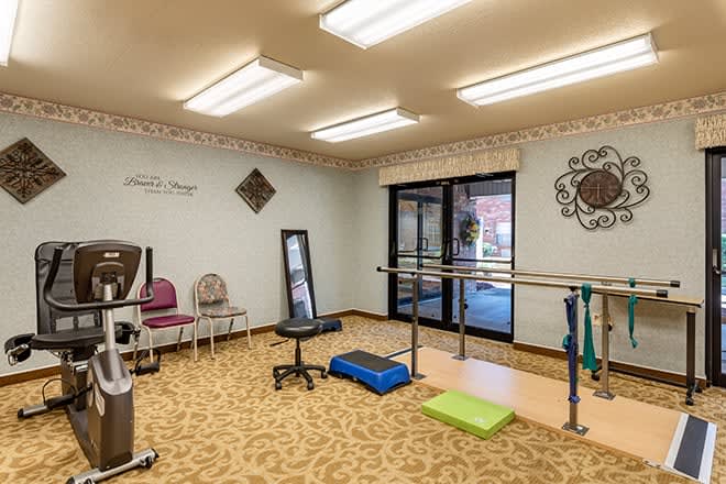 A well-lit physical therapy room equipped with exercise mats, chairs, and parallel bars.