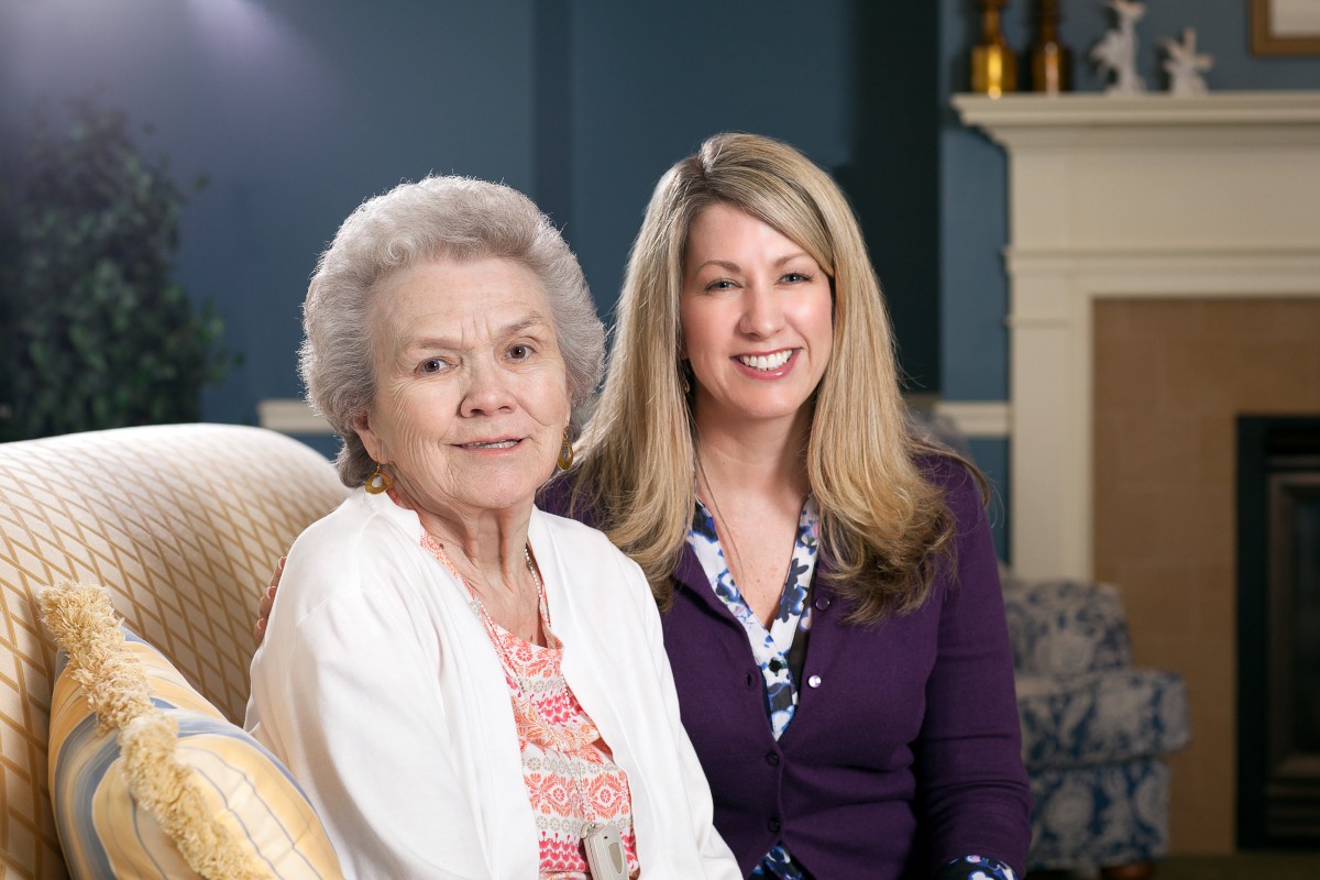 An elderly woman and a younger woman smiling together in a living room.