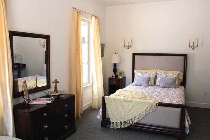 A cozy bedroom featuring a neatly made bed with pillows and a throw blanket, complemented by a dresser with a mirror and soft curtains framing the window.