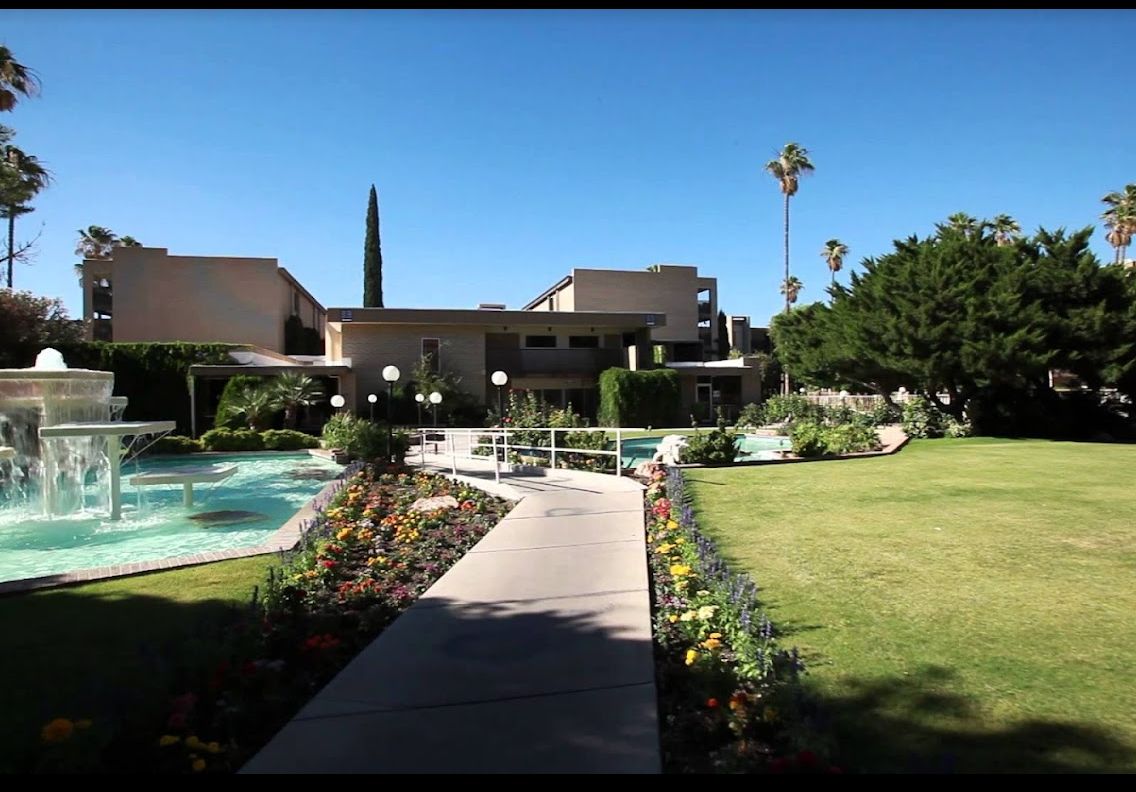 A modern building with a landscaped garden and pathway, surrounded by palm trees under a clear blue sky.