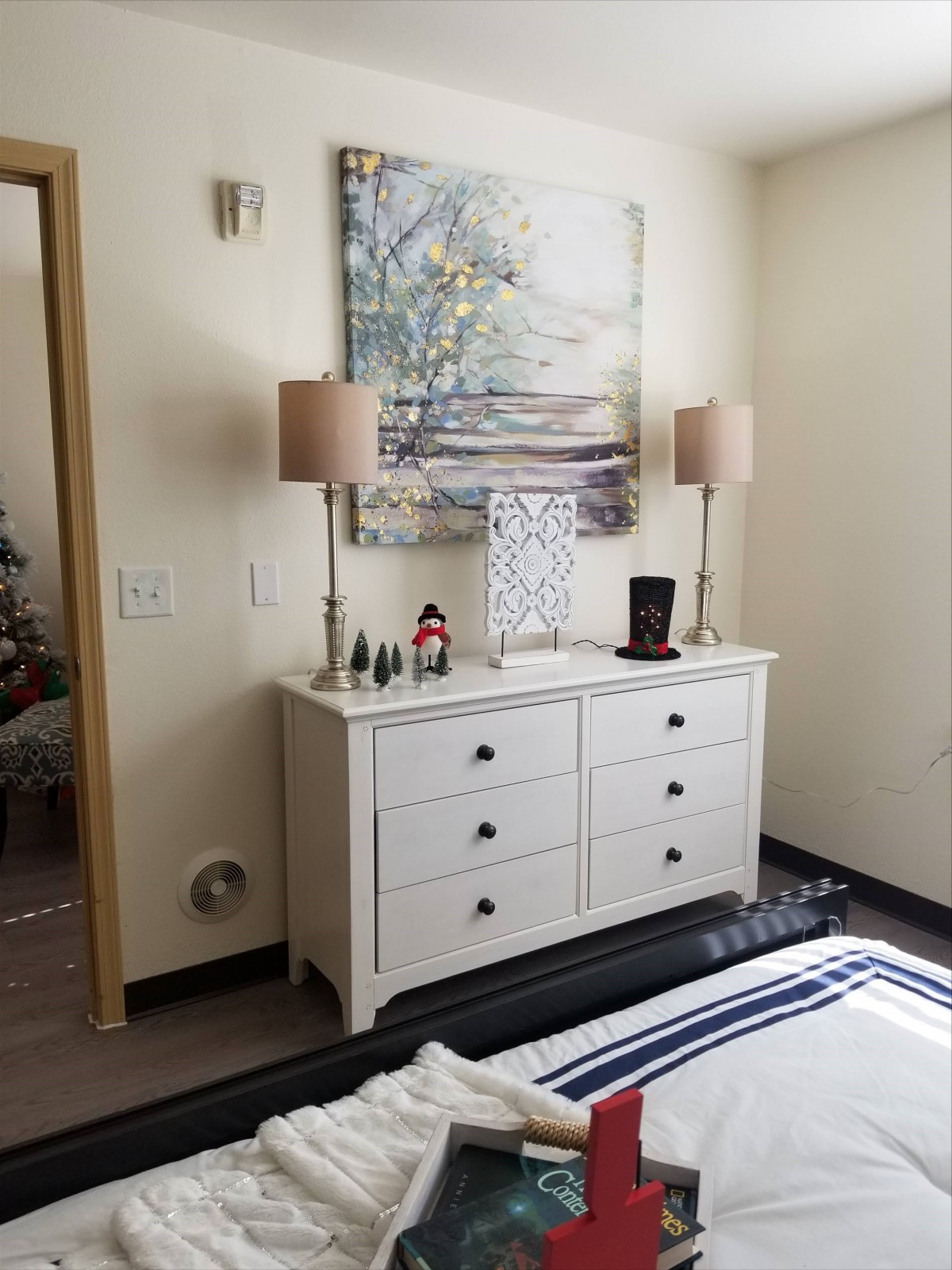 A cozy bedroom with a white dresser, adorned with table lamps and decorative items, and a serene landscape painting on the wall.