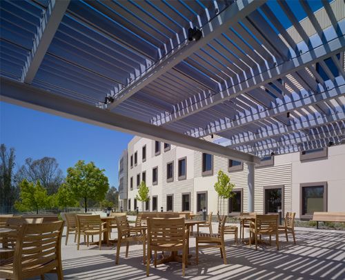 A sunny outdoor patio area with wooden chairs and tables under a modern pergola, adjacent to a contemporary building.