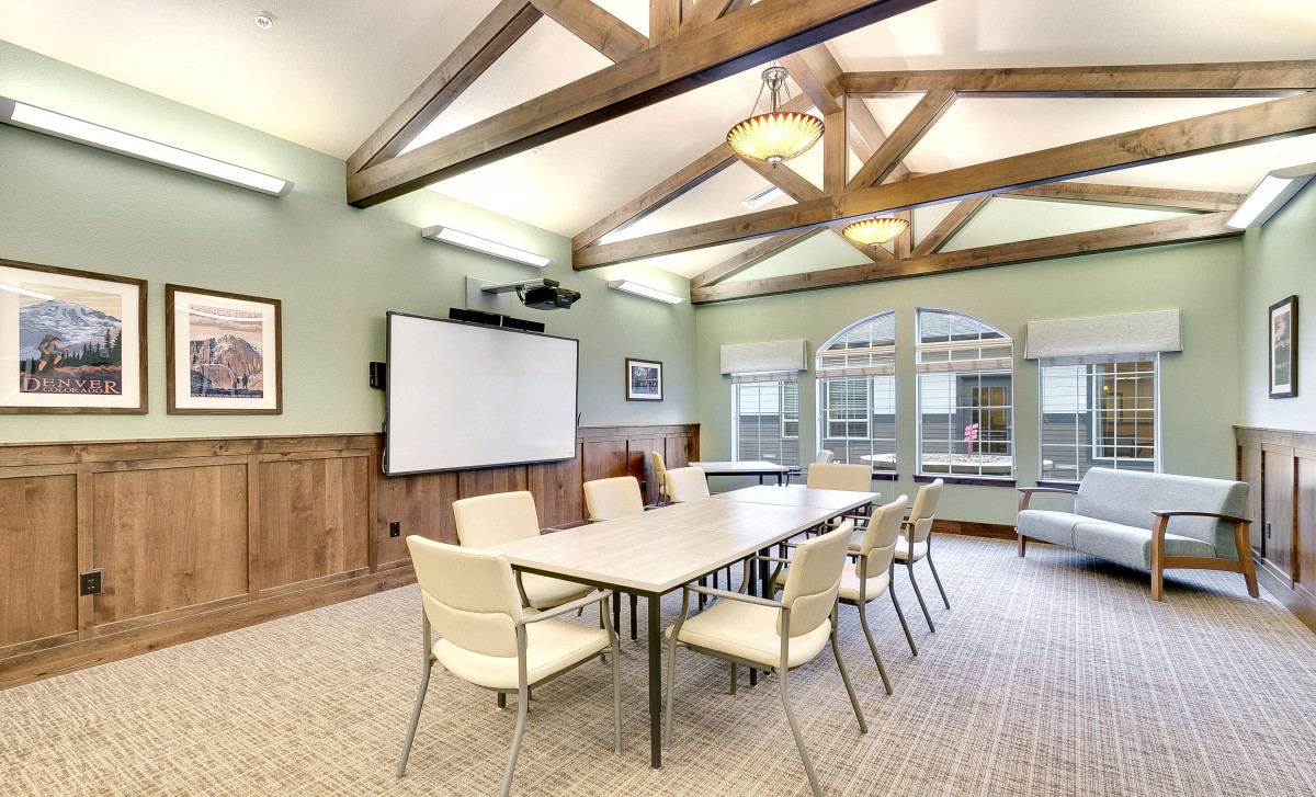 A spacious conference room with a large table, chairs, and a projector screen, highlighted by natural light and wooden ceiling beams.