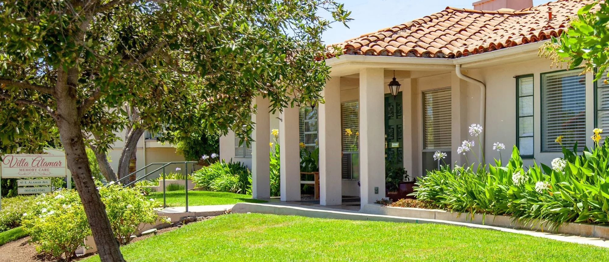 A charming house with a well-maintained lawn and lush greenery surrounding the porch on a sunny day.