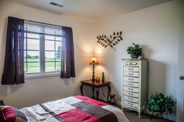 A cozy bedroom with a neatly made bed, a dresser, and a warm lamp on a side table near the window.