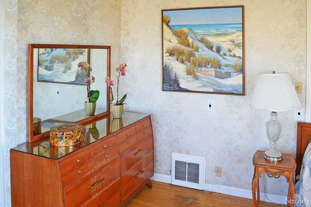 A cozy bedroom corner featuring a wooden dresser with a mirror and decorative items, alongside a landscape painting on the wall.