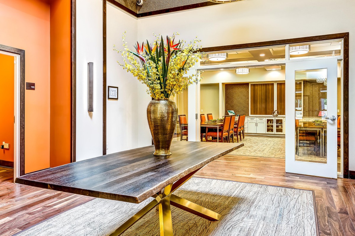 A modern interior space featuring a wooden table with a vase of flowers, leading into a conference room.