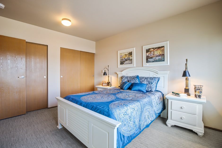 A cozy bedroom featuring a white bed frame with blue bedding, complemented by nightstands and artwork on the walls.