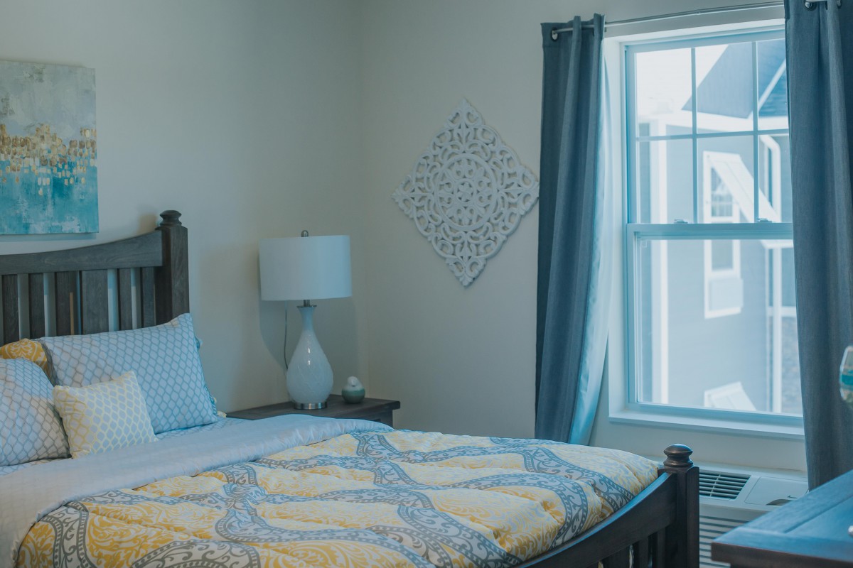 A cozy bedroom with a neatly made bed, accented by a lamp and decorative wall piece near the window.