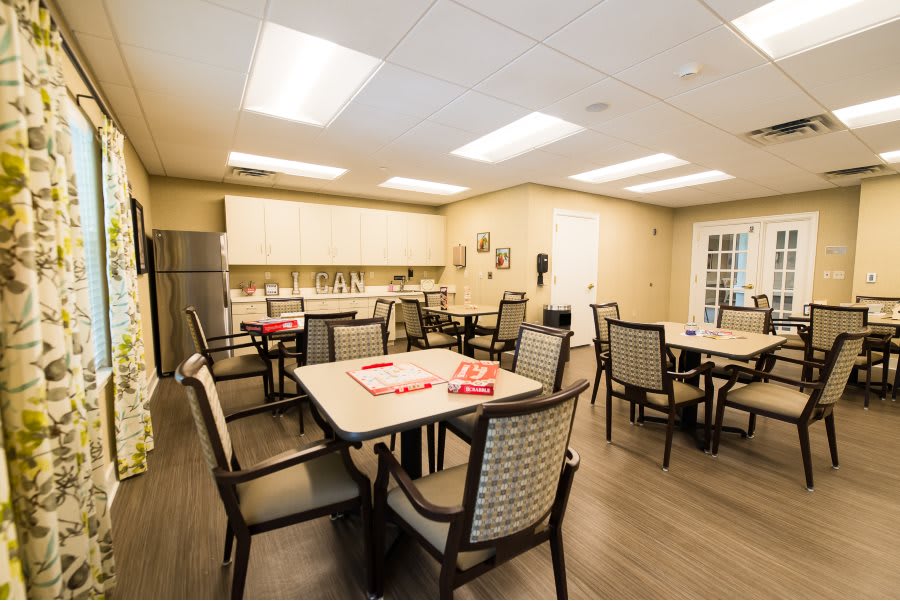 A well-lit dining area with multiple tables and chairs, featuring a kitchenette in the background.
