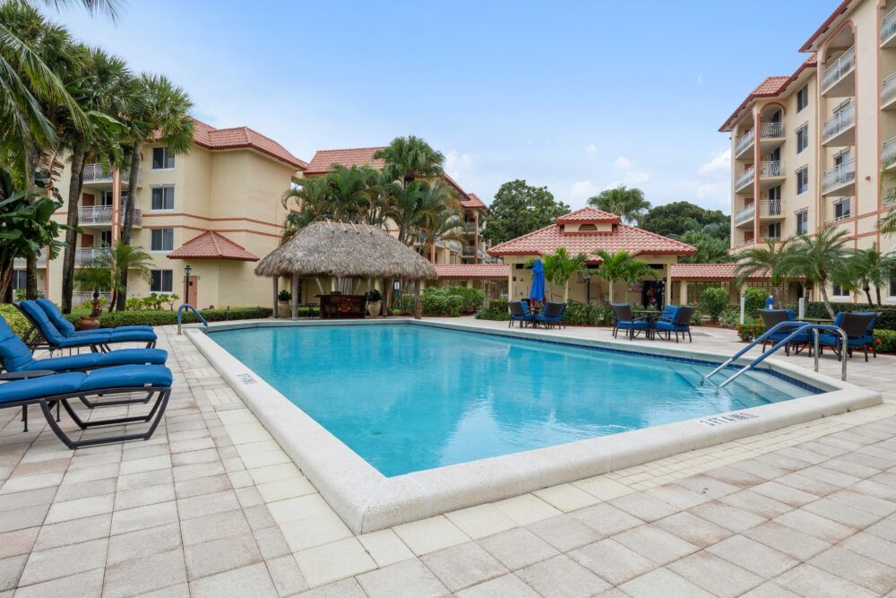 A tropical resort setting featuring a large outdoor swimming pool surrounded by palm trees and residential buildings.