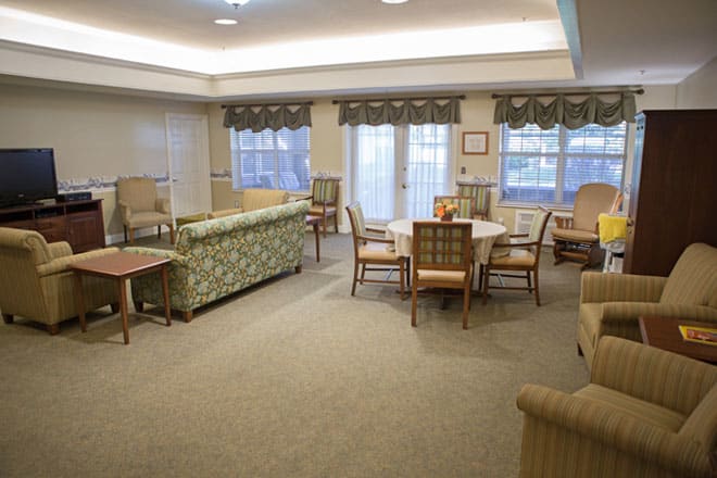 A cozy living room with sofas, chairs, and a table, featuring a carpeted floor and large windows.