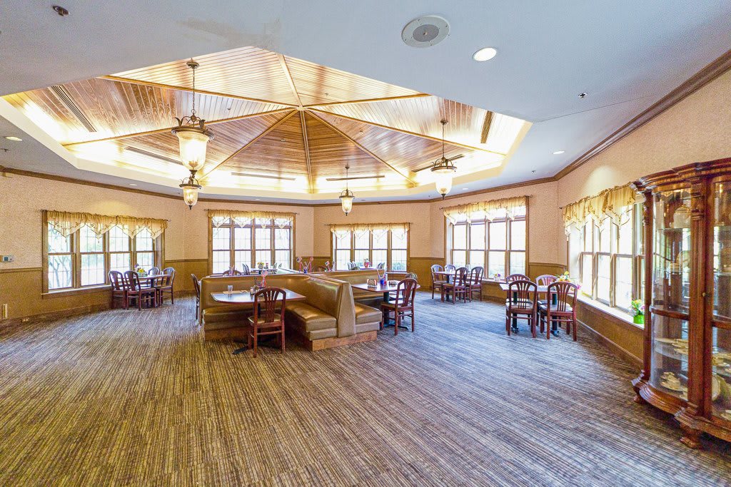 A spacious dining area with wooden floors, tables, and chairs, illuminated by large windows and ceiling lights.