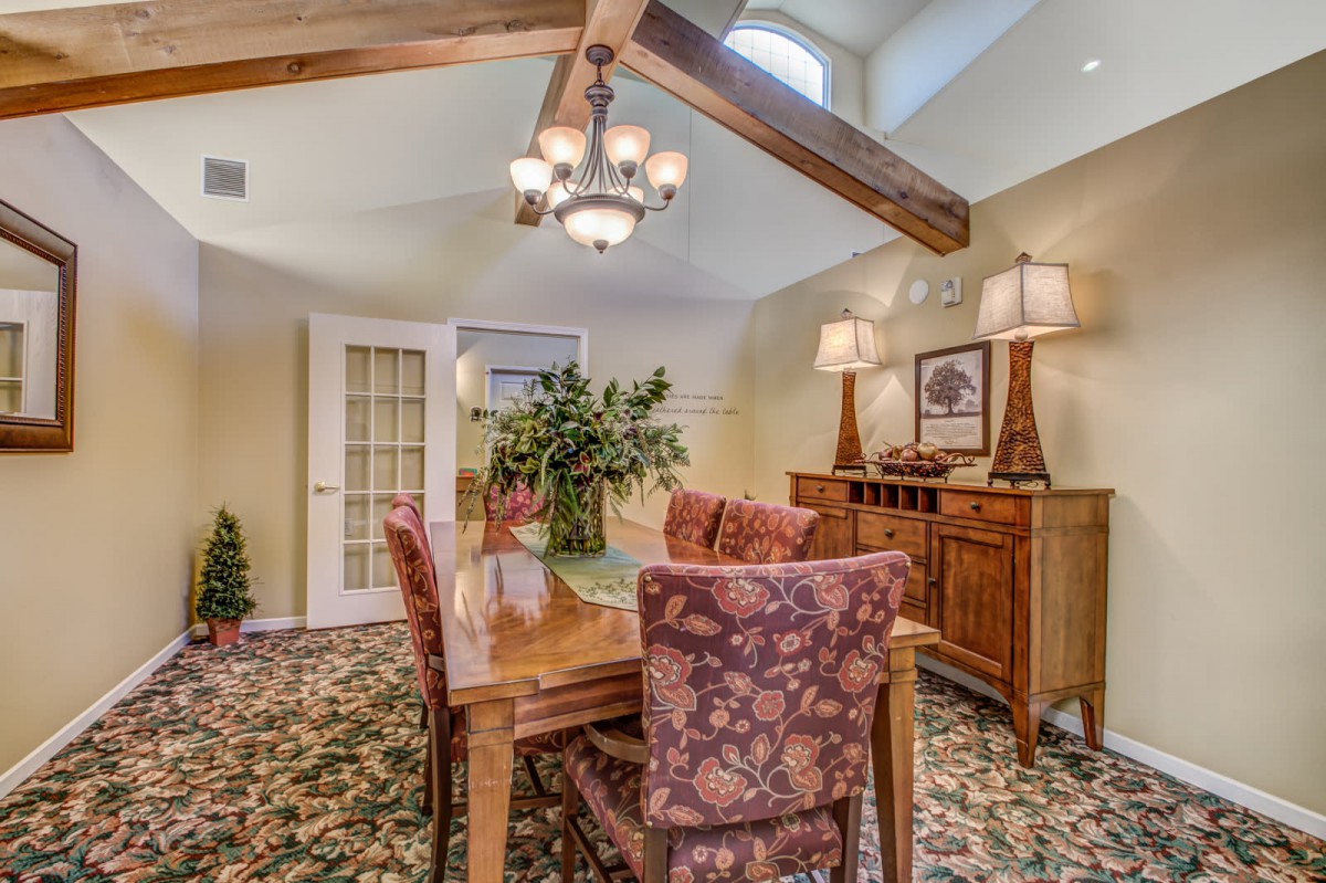 A cozy dining room featuring a wooden table with floral-patterned chairs, illuminated by a chandelier and complemented by decorative lamps and plants.