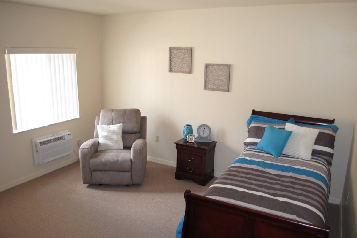A cozy bedroom with a bed, armchair, and nightstand, featuring neutral tones and colorful accents.